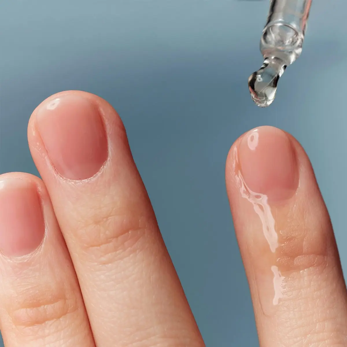 Lähikuva sormista, joille levitetään läpinäkyvää ihonhoitoseerumia pipetillä vaaleansinistä taustaa vasten. Kosteuttava skincare-seerumi korostuu kiiltävällä pinnalla ja minimalistisella beauty-ilmeellä.