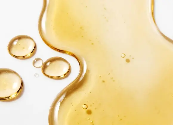 Närbild av gyllene ansiktsolja eller serum i flytande form med droppar och mjuka former på en ljus bakgrund, med fokus på hudvård och återfuktande serum.