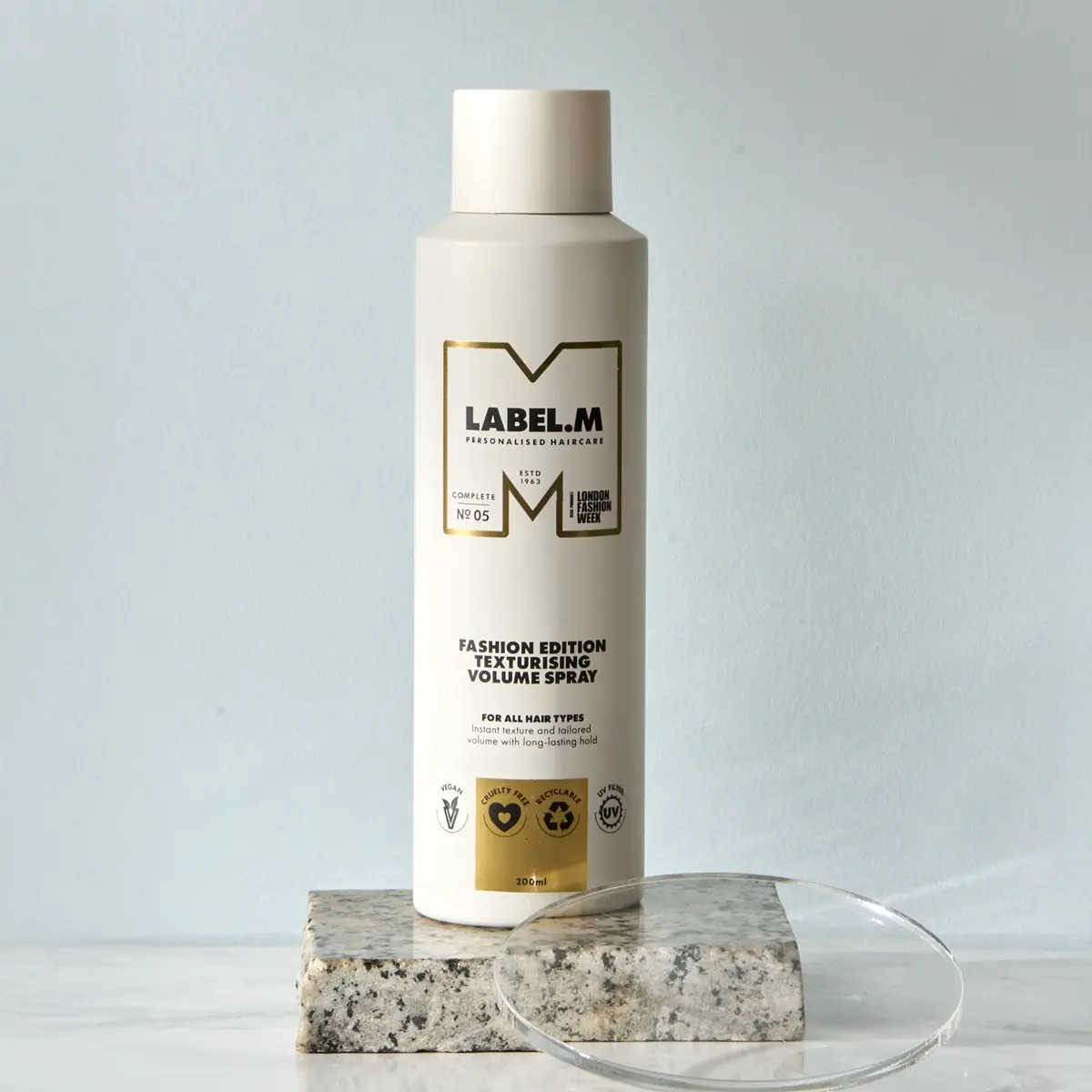 LABEL.M Fashion Edition Texturising Volume Spray i vit flaska placerad på ett stenblock med en transparent akrylskiva framför. Förpackningen visar texten “LABEL.M Personalised Haircare”, “Fashion Edition Texturising Volume Spray”, “For All Hair Types” och volymen 200 ml.