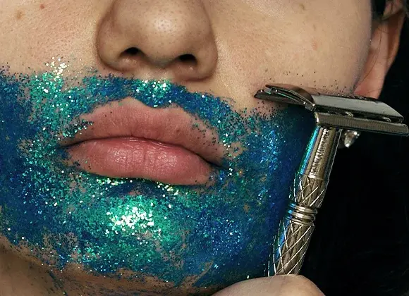 Lähikuva suusta ja leuasta, iholla sinivihreää glitteriä, ja metallinen partahöylä koskettaa leukaa tummalla taustalla.