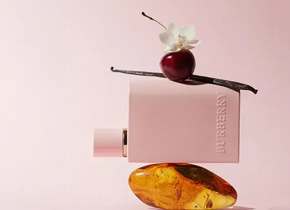 En rektangulær, lyserosa flaske med teksten «BURBERRY» liggende horisontalt. Flasken balanserer på en ravfarget stein. Oppå flasken ligger en mørk vaniljestang, et rødt bær og en hvit blomst. Bakgrunnen er ensfarget i en myk rosa tone.