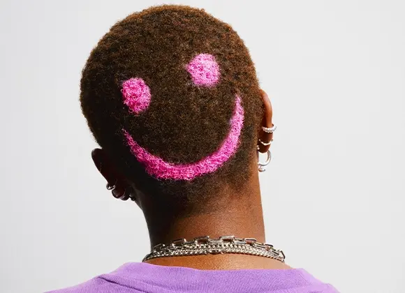 Bakhuvud med kort, lockigt hår där ett rosa smiley-motiv är infärgat i håret. Personen bär lila tröja, flera örhängen och ett silverfärgat halsband.