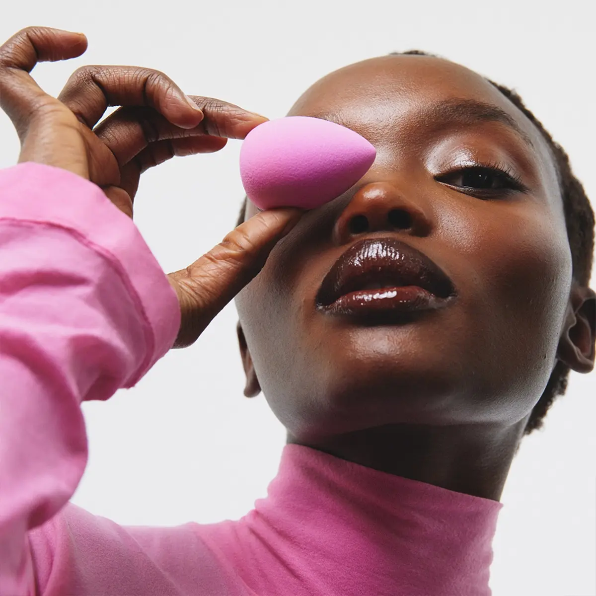 En person i rosa polotröja håller en rosa makeupsvamp mot området kring ögat. Närbild med glansig makeup på läppar och ögon.