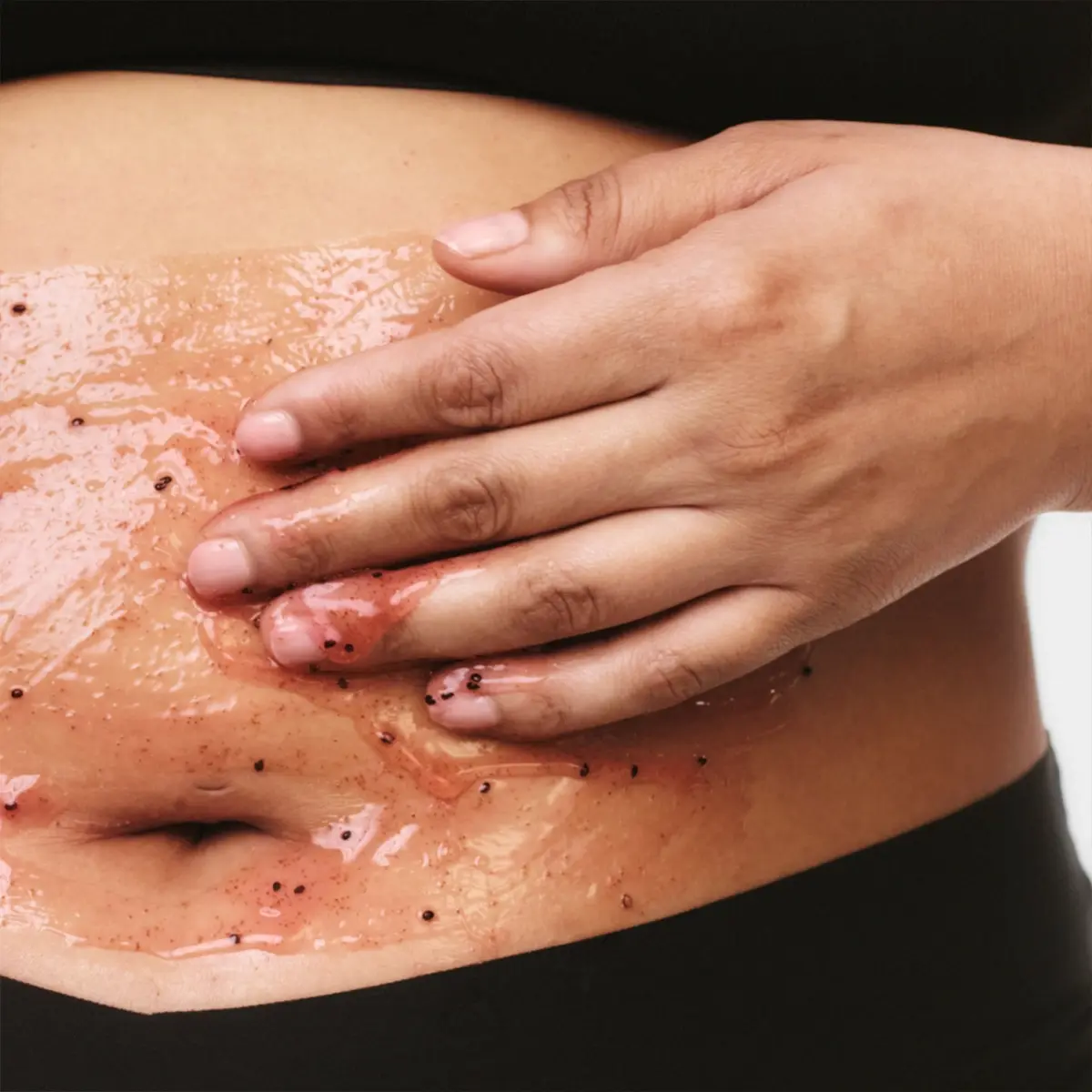 En hand applicerar kroppsskrubb med synliga exfolierande korn på magen. Bilden visar kroppsvård och exfoliering med gelbaserad scrub på huden.