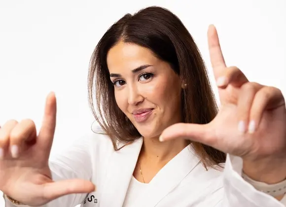 En person i vit arbetsrock håller upp händerna framför kameran och formar en rektangel med fingrarna som en kameraram. Närbild med naturlig makeup, neutral bakgrund och fokus på ansiktet och händerna.