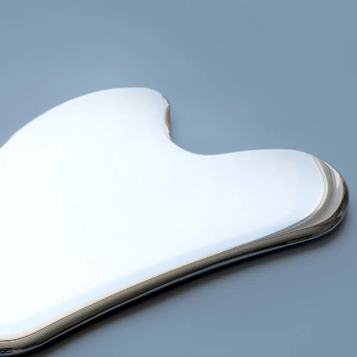 Kiiltävä, hopeanvärinen metallinen gua sha, jossa on pehmeästi kaartuvat reunat, harmaansinistä taustaa vasten.