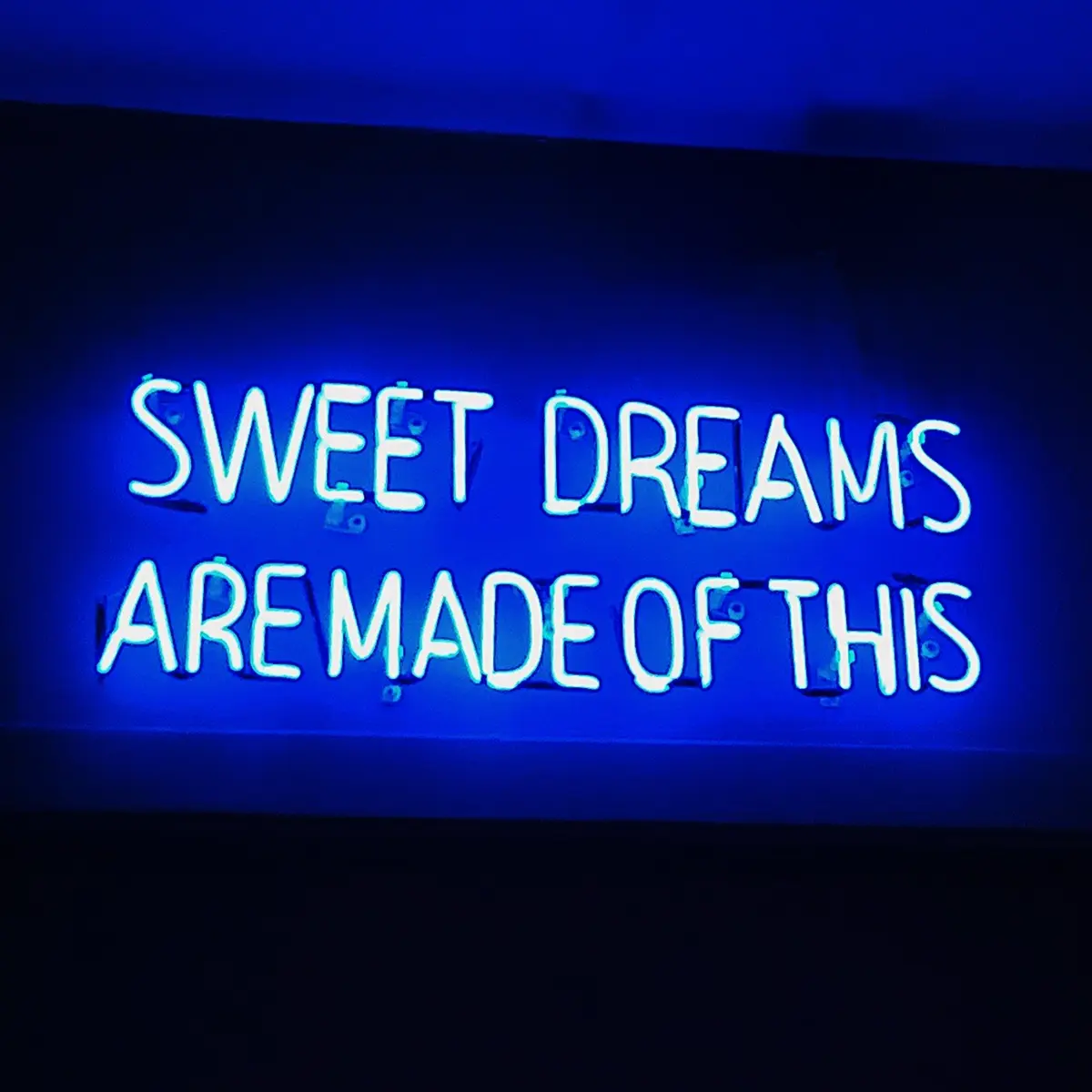 Blå neonskylt på en mörk vägg med den lysande texten “SWEET DREAMS ARE MADE OF THIS”. Skylten ger ett blått sken mot omgivningen och är bildens centrala motiv.