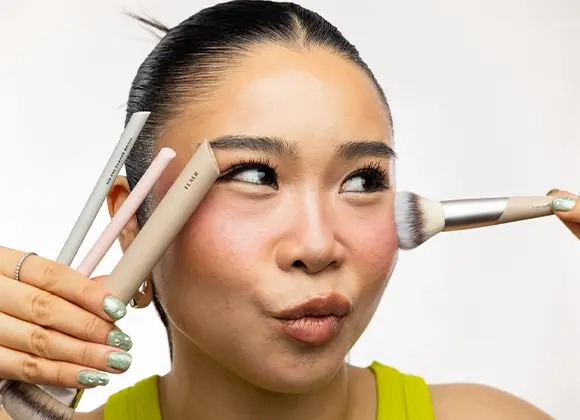 Person med mörkt hår uppsatt i låg knut applicerar highlighter med sminkborste på kindbenet. Personen håller flera beige och ljusa sminkborstar i ena handen.