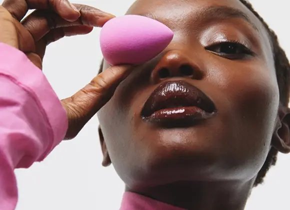 Närbild av en person i rosa polotröja mot ljus bakgrund. Personen håller en rosa sminksvamp framför det ena ögat med handen. Ansiktet är lätt vinklat uppåt och läpparna har blank makeup.