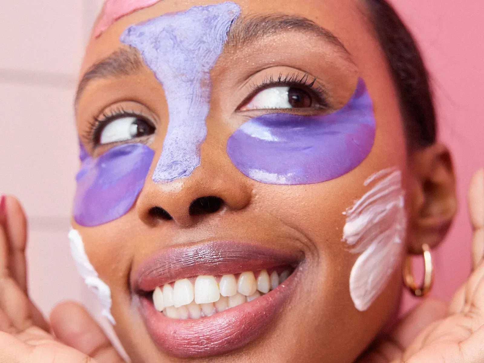 Leende kvinna med färgglada ansiktsmasker i lila och vitt, samt ögonpads, framför rosa bakgrund