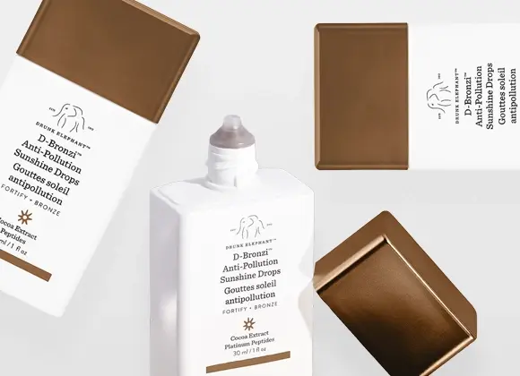 Tuotekuva, jossa useita Drunk Elephant D-Bronzi Anti-Pollution Sunshine Drops -pulloja ja korkkeja. Valkoisia, neliskulmaisia pulloja pronssinvärisillä korkeilla eri asennoissa vaaleaa taustaa vasten.