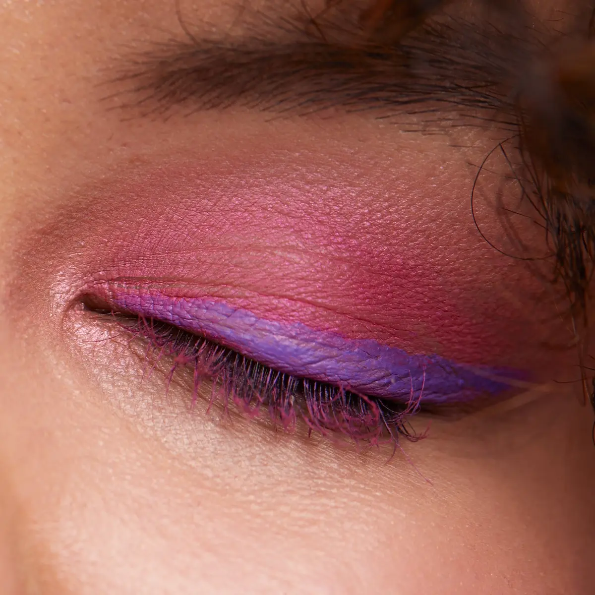 Närbild av ögonmakeup med rosa ögonskugga och lila eyeliner på slutna ögonlock, samt rosa mascara på ögonfransarna.
