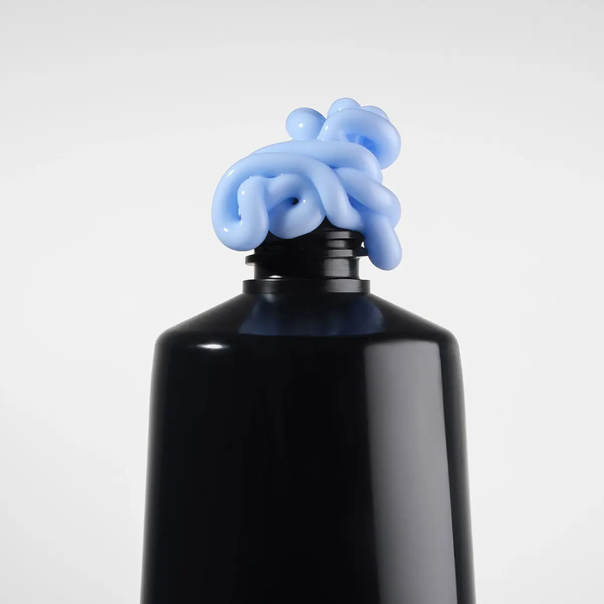 Närbild av en svart kosmetikflaska med ljusblå hudvårdskräm som ringlar ut ur den öppna flaskhalsen mot en ljus, enfärgad bakgrund. Fokus ligger på produktens krämiga konsistens och minimalistiska förpackning.