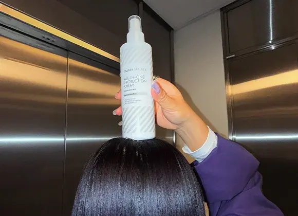 Käsi pitelee Matas Striber All-In-One Protection Spray -pulloa kiiltävän, tumman ja sileän hiuksen yläpuolella hississä.