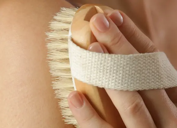 Närbild på en torrborste med trähandtag och ljusa naturborst som används mot huden för kroppspeeling och exfoliering. En hand håller borsten med vit textilrem medan den borstar huden i en hudvårdsrutin för body care.