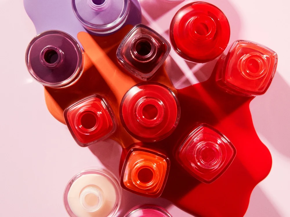 Useita avonaisia Atelier Rouge -kynsilakkapulloja ylhäältä kuvattuna, väriläiskiä punaisena, oranssina ja violettina vaaleanpunaisella taustalla.