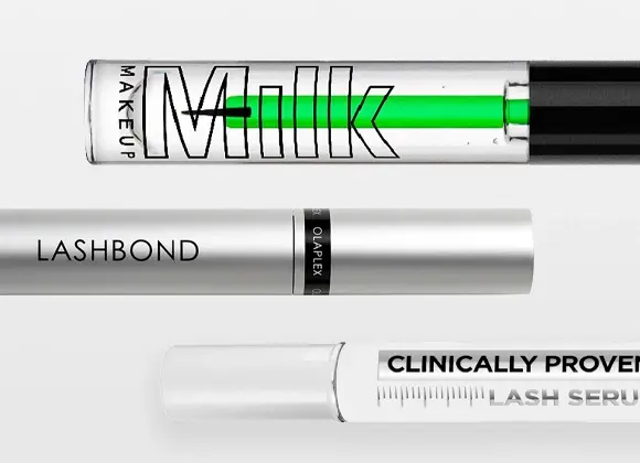Lähikuva ripsiseerumista ja ripsiväristä merkeiltä Olaplex ja Milk Makeup; pakkauksissa tekstit "Milk Makeup", "LASHBOND", "OLAPLEX" ja "CLINICALLY PROVEN LASH SERUM" läpinäkyvässä ja valkoisessa designissa.