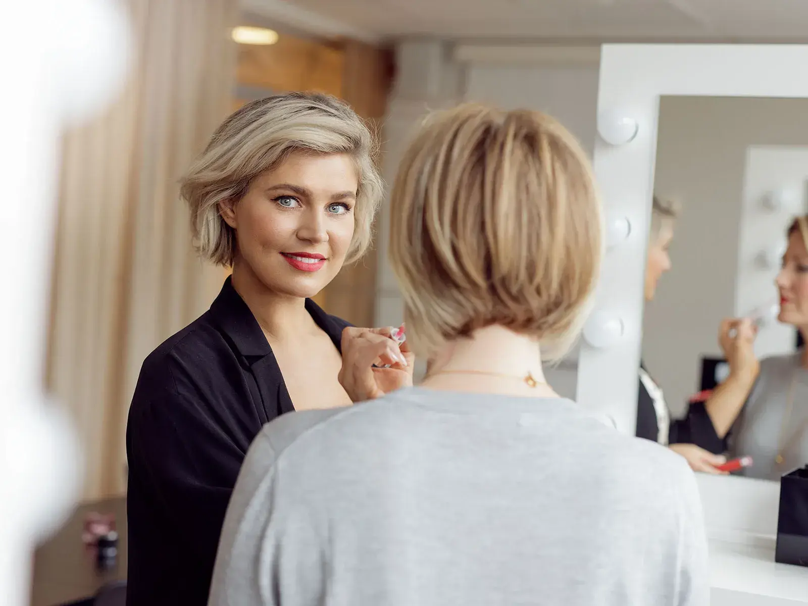 Blond kvinna i fokus applicerar läppstift på en annan kvinna med ryggen mot kameran, i ett rum med beiga gardiner, vita väggar och en stor spegel.