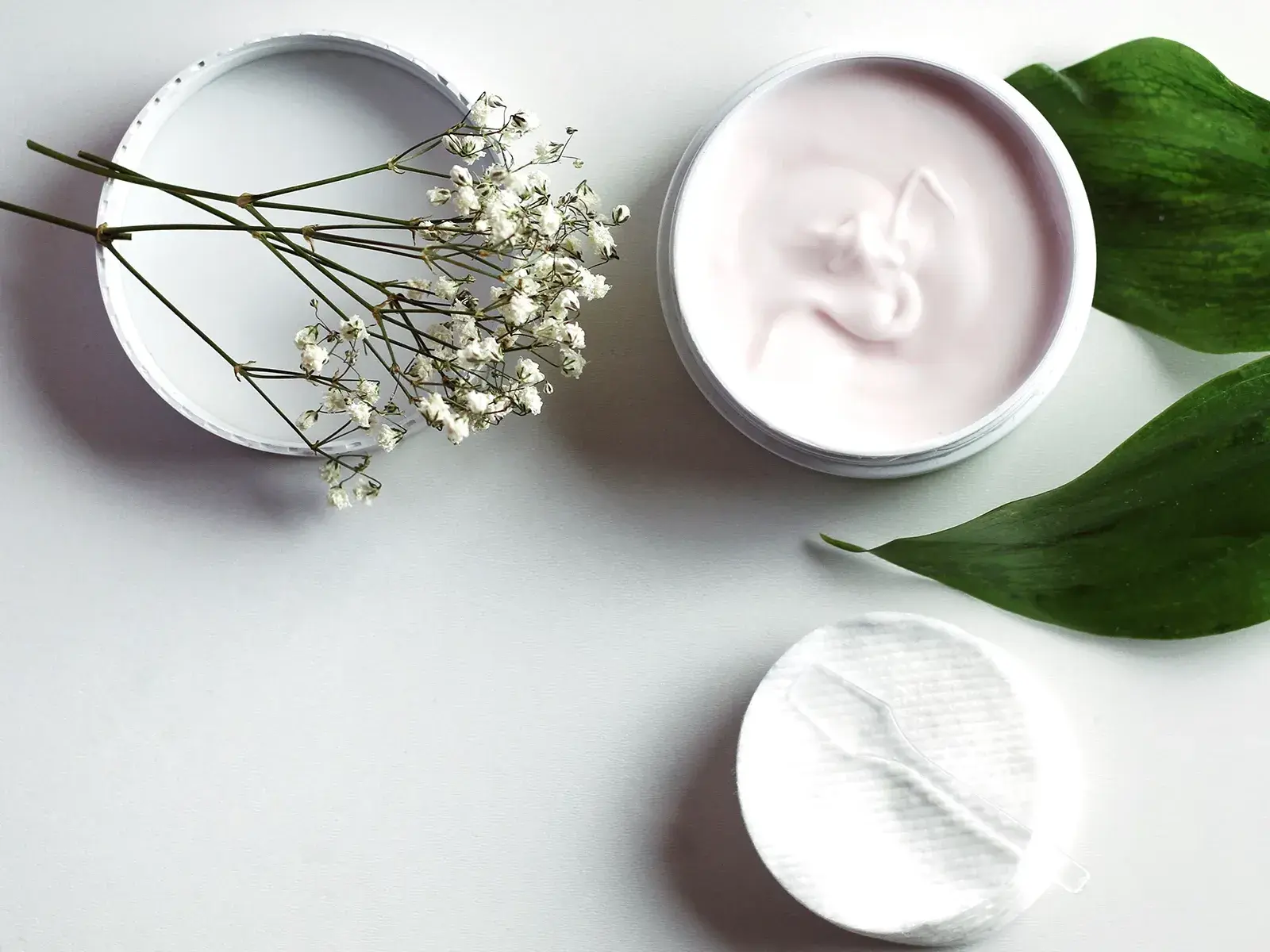 Avoin purkki, jossa on vaaleanpunaista voidetta, ympäröitynä vihreillä lehdillä ja pienillä valkoisilla kukilla.