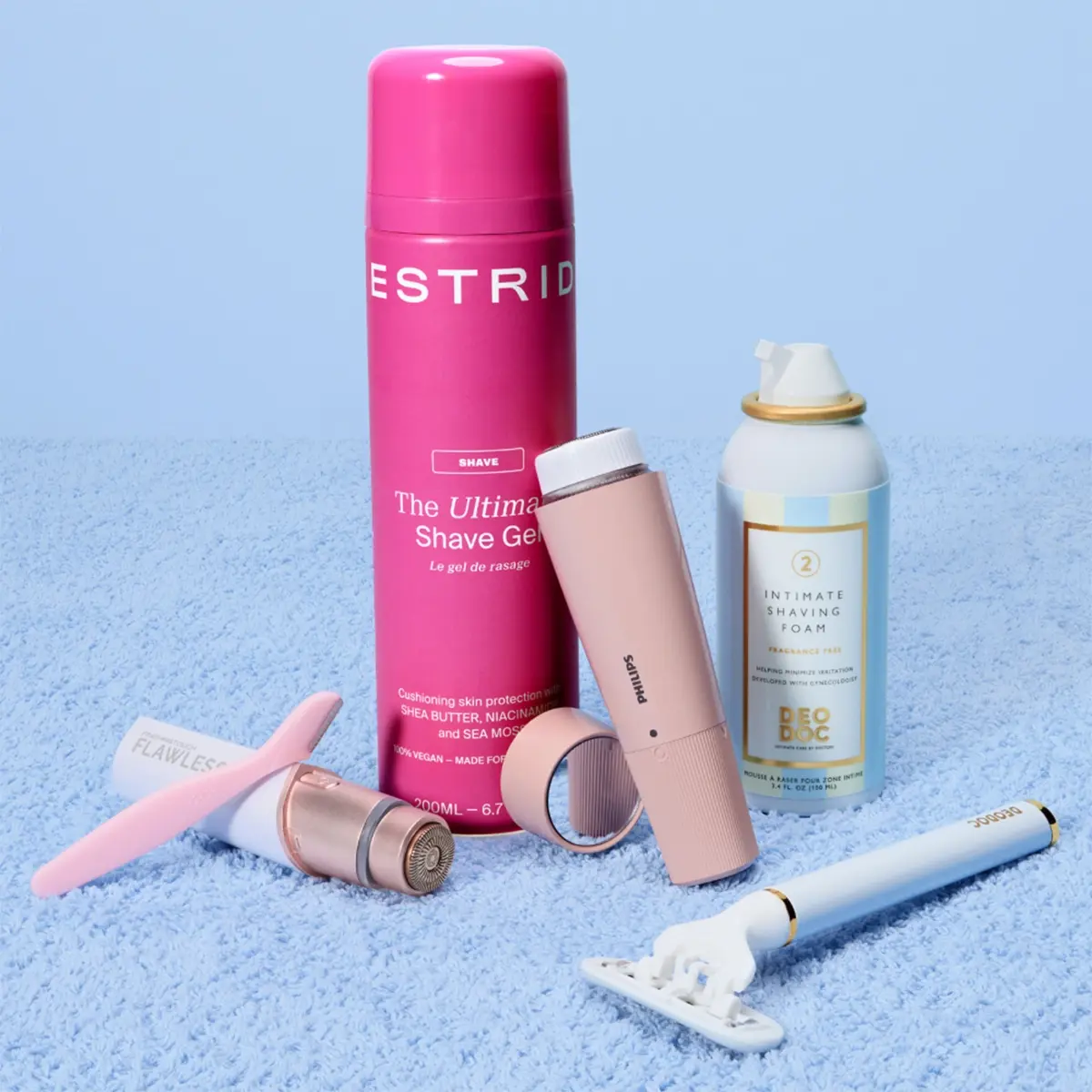 Sheivaus- ja intiimihygieniatuotteita esillä vaaleansinistä taustaa vasten, mukana muun muassa Estrid The Ultimate Shave Gel, DeoDoc Intimate Shaving Foam Fragrance Free, Philips sähkötrimmeri ja Flawless-karvanpoistolaite. Tuotteet on esitetty vaaleanpunaisissa, valkoisissa ja pastellisävyisissä pakkauksissa yhdessä höylän ja grooming-välineiden kanssa intiimialueen sheivaukseen ja ihonhoitoon.