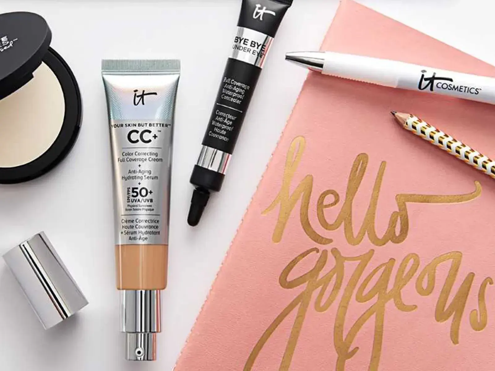 Flere produkter fra IT Cosmetics plassert på en hvit overflate, sammen med en rosa duk med teksten "hello gorgeous" i gull.