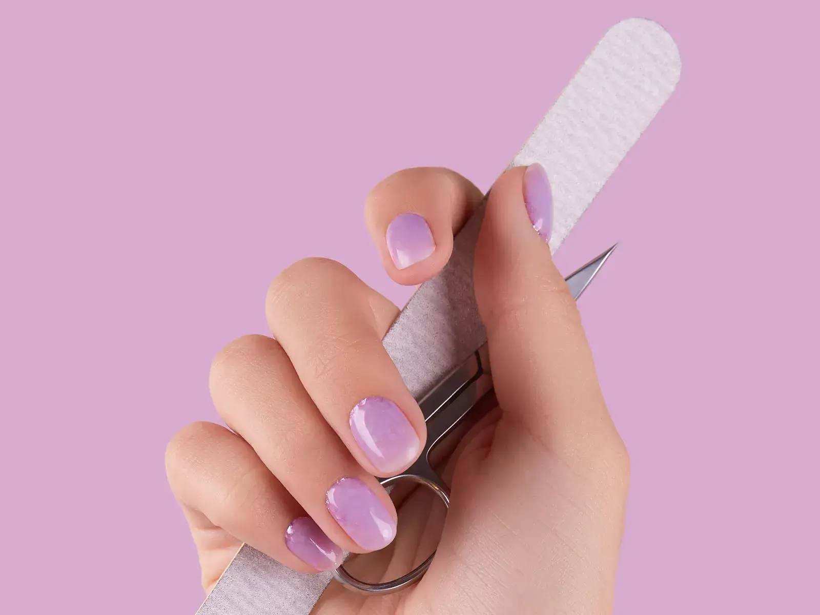 Hand som håller en nagefil och en nagelsax. Naglarna är lila och bakrgunden lila. 