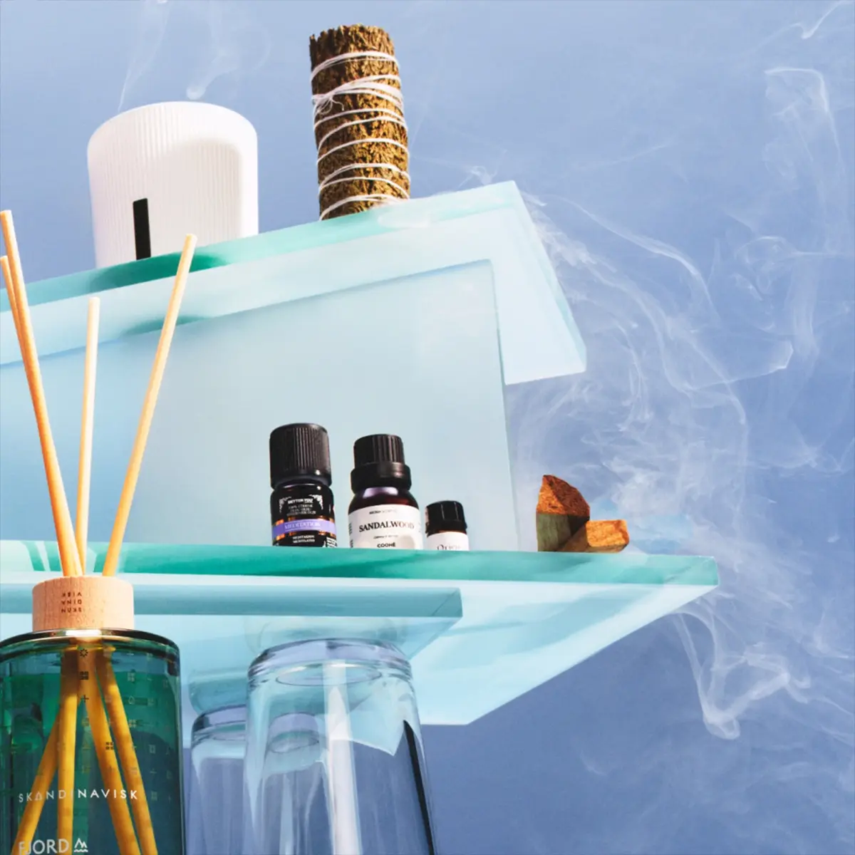 Tuoksutuotteita ja aromaterapiaa läpinäkyvillä lasitasoilla sinistä taustaa vasten savuefekteillä. SKANDINAVISK FJORD -huonetuoksu tuoksutikuilla on aseteltu eteeristen öljyjen, kuten “Sandalwood”, valkoisen diffuserin ja salvianipun viereen kodin tuoksumaailmaa ja wellness-tunnelmaa varten.
