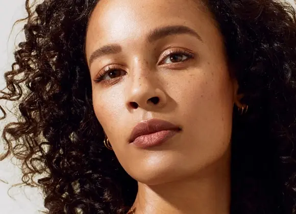 Porträtt av en person med naturlig makeup, i närbild och halvkroppsbild, med guldaccessoarer mot ljus bakgrund.