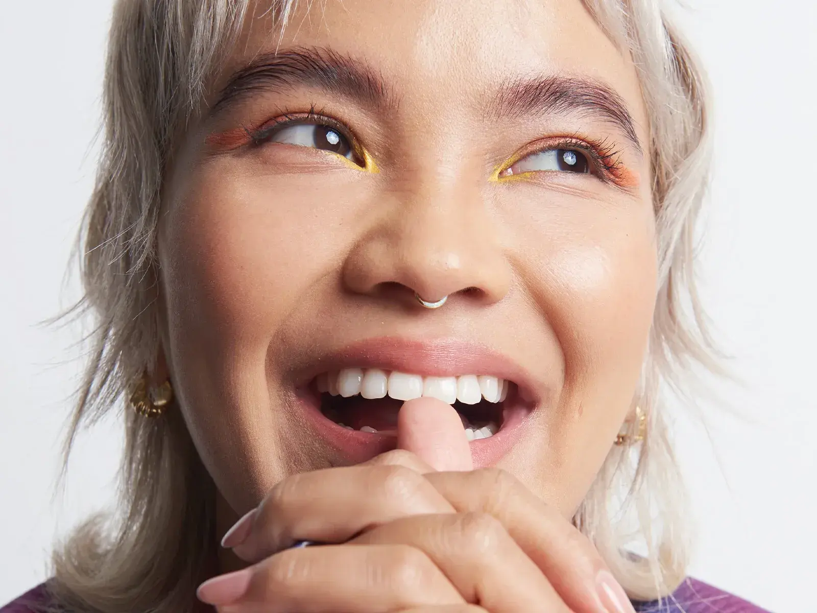Kvinna med makeup, näspiercing och kort, blondt hår, som ler med öppen mun, tittar åt sidan och har händerna knutna framför hakan mot en vit bakgrund.