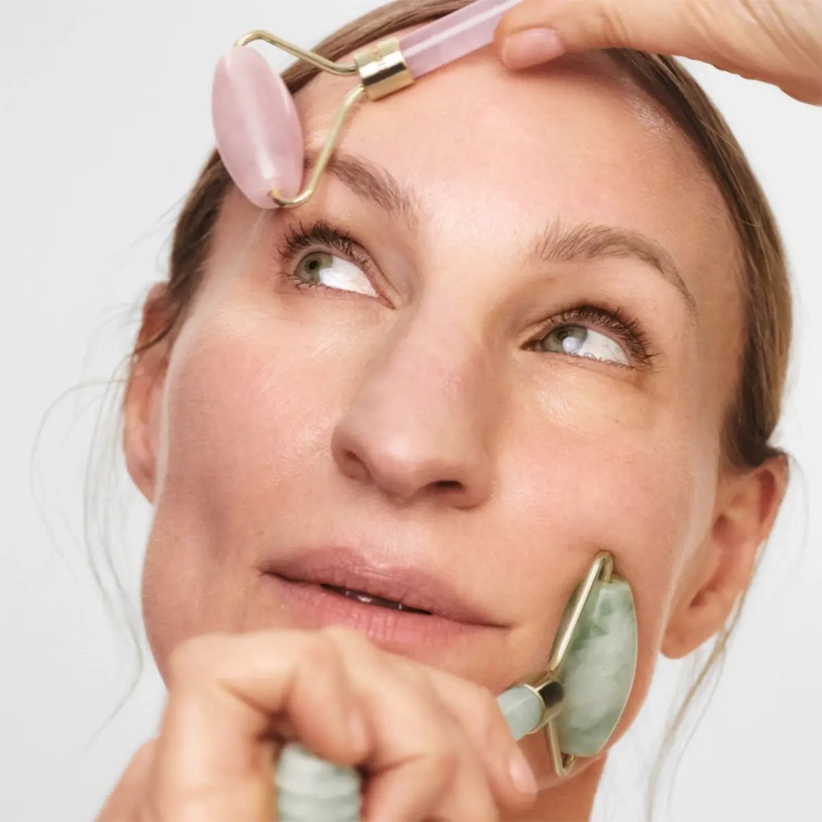 Lähikuva henkilöstä, joka käyttää kahta vaaleanpunaista ja vihreää kivistä valmistettua kasvorullaa otsalla ja poskella osana skincare-rutiinia. Ihonhoitotyökalu kasvohierontaan ja glow-efektiin vaaleaa taustaa vasten.