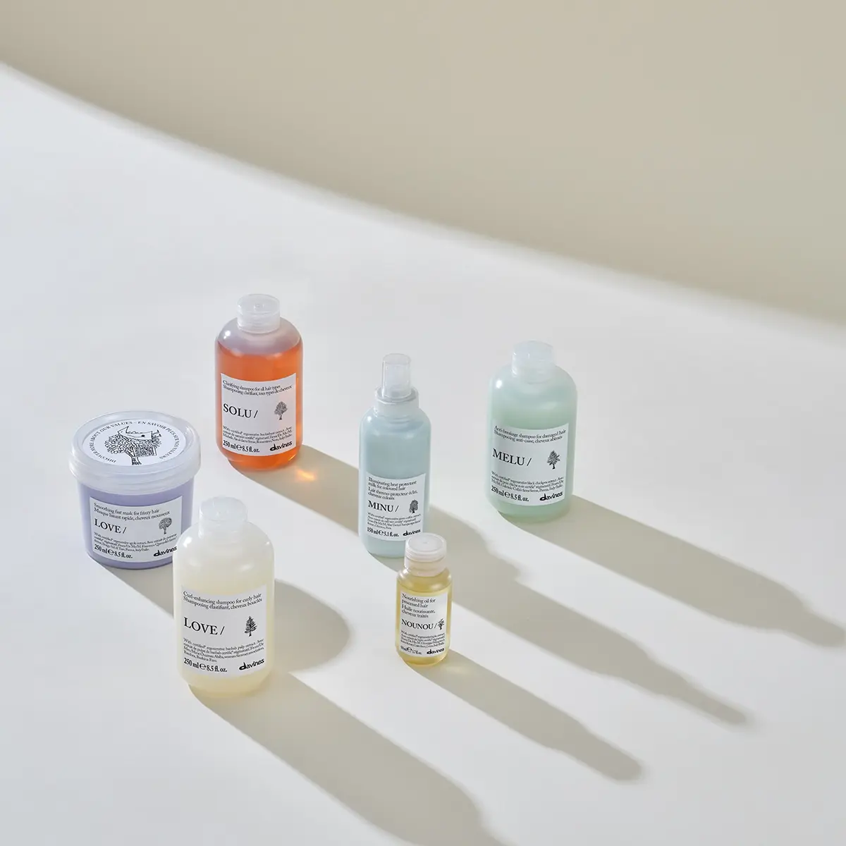 Davines hårvårdsprodukter från serierna LOVE, SOLU, MINU, MELU och NOUNOU står uppställda på vit yta med långa skuggor i mjukt ljus. Bilden visar schampo, hårmask, hårolja och värmeskyddsspray med produktnamn och Davines-logotyp synliga på förpackningarna.