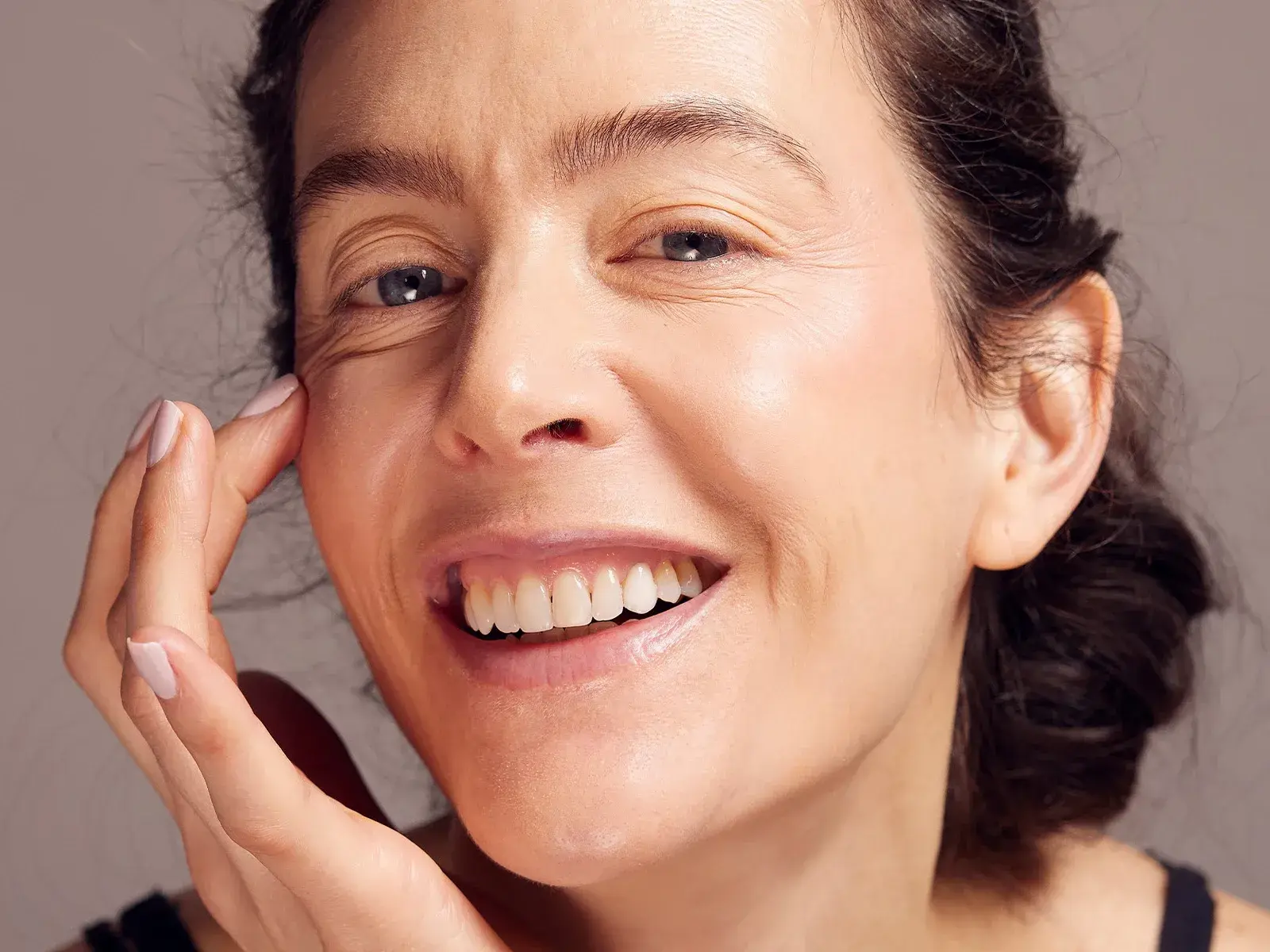 Närbild på en leende kvinna med bar hud och naturlig makeup som försiktigt rör vid huden under ena ögat.