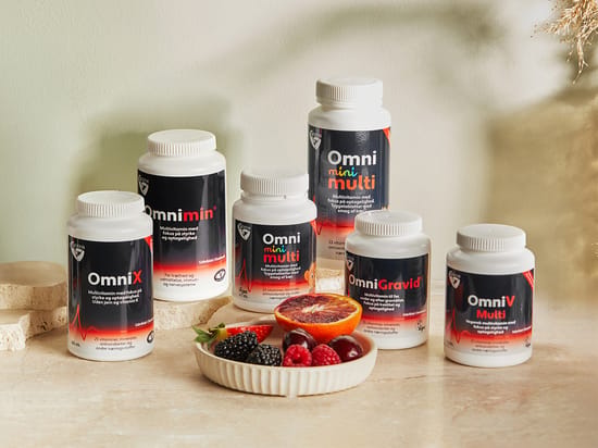Seks forskellige Omni-vitaminprodukter fra Biosym står på en lys beige overflade med bær og en halv blodappelsin foran. Varmt sollys skaber skygger i baggrunden.