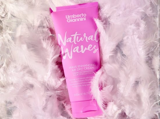 Her ses Umberto Giannini Natural Waves Wave Enhancing Air Dry Cream i en pink tube, omgivet af lyserosa og hvide fjer. På emballagen står blandt andet “Natural Waves”, “Wave Enhancing Air Dry Cream” og “200 ml”.