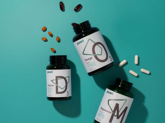 Tre Puori-kosttilskud med D3-vitamin, Omega 3 og magnesium ligger på en turkis baggrund omgivet af forskellige kapsler og tabletter.