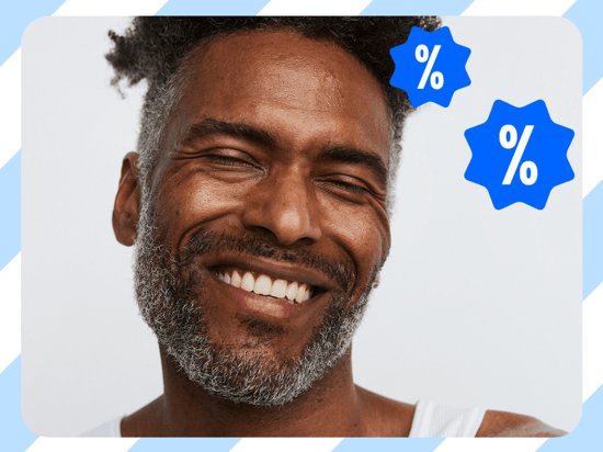 Her ses en smilende person med kort, mørkt hår og gråt skæg mod en lys baggrund. Øverst til højre ses to blå, takkede symboler med hvide procenttegn (%).