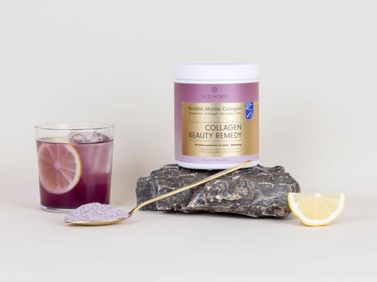 Vild Nord Collagen Beauty Remedy på en mørk sten ved siden af et glas lilla drik med citron, en ske med pulver og en citronskive.