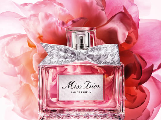 Her ses Miss Dior Eau de Parfum i en transparent flakon med rosa indhold og en sølvfarvet sløjfe. Flakonen står foran store blomster i rosa og pink nuancer.