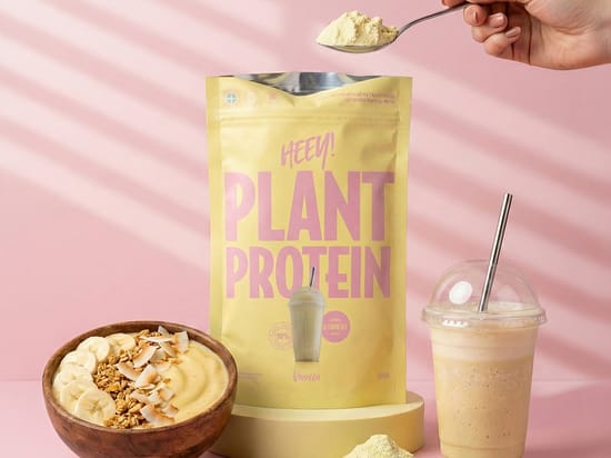 En gul pose Heey! Plant Protein med vaniljesmag står centralt på en gul sokkel mod en lyserød baggrund. Foran ses en skål med smoothie, bananskiver, granola og kokos samt et glas proteinshake, mens en hånd holder en ske med pulver over posen.