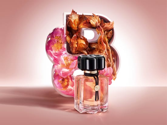 Her ses BOSS Bottled Beyond for Her i en transparent parfumeflakon med rosa indhold, sort låg og guldfarvede detaljer. Bag flakonen ses et stort BOSS-logo dekoreret med rosa blomster og brune blade mod en lys rosa baggrund.
