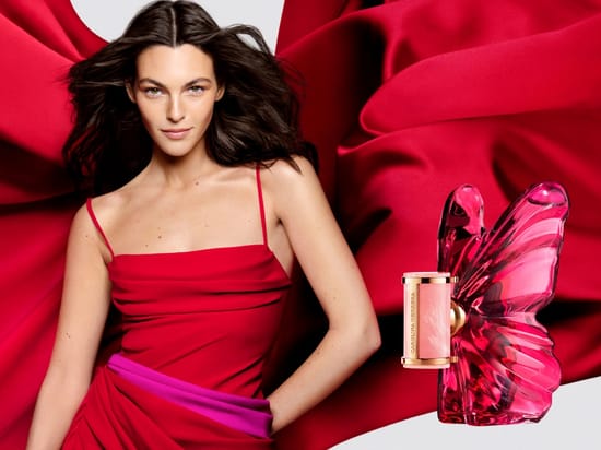 Her ses et billede af en person iført en rød kjole foran en baggrund med rødt stof. Til højre ses en lyserød parfumeflakon fra Carolina Herrera formet som en sommerfugl.