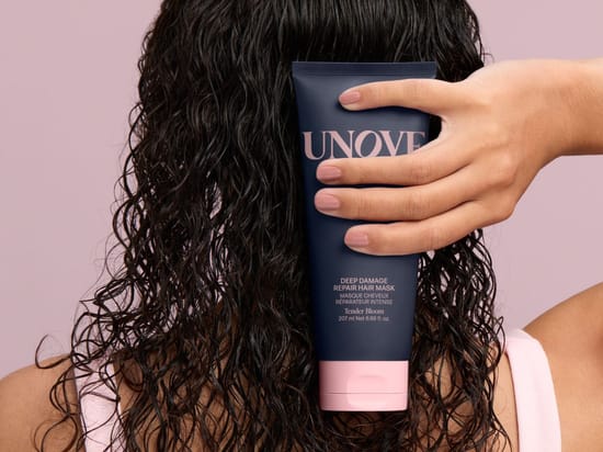 Her ses en person med langt, vådt, mørkt og krøllet hår, der holder UNOVE Deep Damage Repair Hair Mask foran håret. På emballagen står blandt andet “Deep Damage Repair Hair Mask”, “Tender Bloom” og “207 ml”