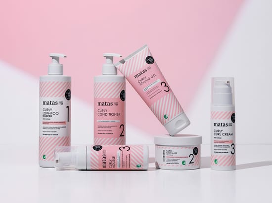 Her ses et billede med produkter fra Matas Stribers Curly-serie placeret mod en lyserød baggrund. Serien omfatter blandt andet Curly Low-Poo Shampoo, Curly Conditioner, Curly Styling Gel, Curly Hair Mask, Curly Curl Cream og Curly Mousse. Produkterne er arrangeret samlet, og den lyserøde emballage med hvide striber gør det tydeligt, at de tilhører samme produktserie.