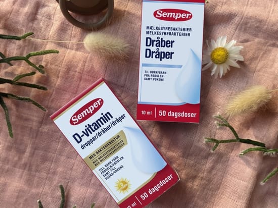 To pakker Semper dråber på rosa stof med blomster omkring. Produkterne er D-vitamin dråber og mælkesyrebakteriedråber, begge med 50 dagsdoser.