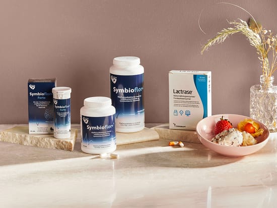 Flere Symbioflor-produkter og en pakke Lactrase står på en lys stenoverflade foran en varm brun baggrund. Til højre står en lyserød skål med yoghurt, jordbær og physalis samt en lille vase med tørrede strå.