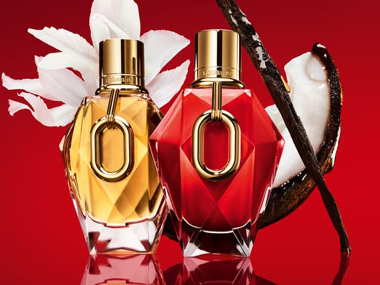 Her ses to Rabanne Million-parfumeflakoner i gyldne og røde nuancer med guldfarvede detaljer. Bag flakonerne ses en hvid blomst, en åbnet kokosnød og vaniljestænger mod en rød baggrund.
