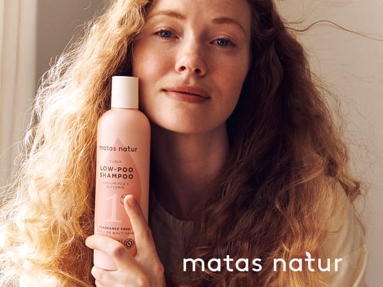 Her ses en person med langt, bølget hår, der holder Matas Natur Curly Low-Poo Shampoo.