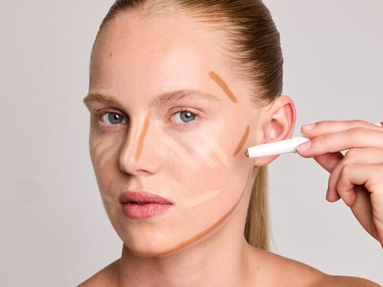 Nærbillede af model med makeupstreger i flere nuancer på ansigtet, mens en GOSH Melt Stick påføres på kinden.