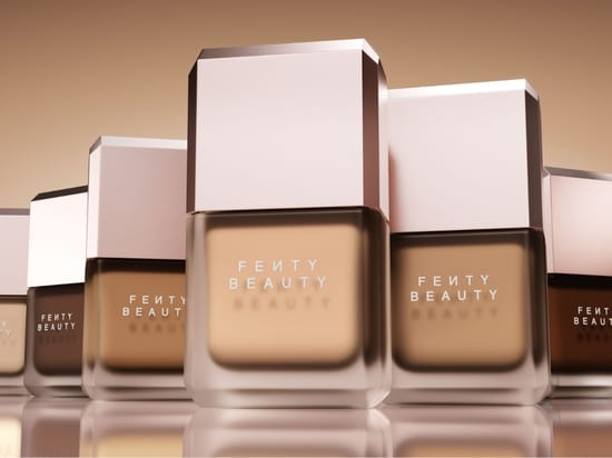 Her ses flere Fenty Beauty foundationflasker i forskellige nuancer med lyse metalliske låg. Flaskerne er placeret tæt sammen mod en varm beige baggrund.