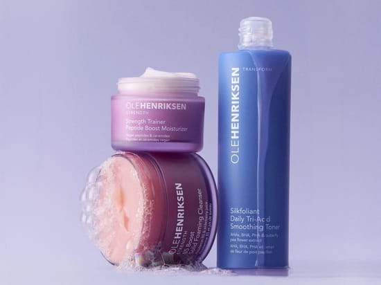 Her ses tre hudplejeprodukter fra Ole Henriksen: Strength Trainer Peptide Boost Moisturizer, Truth Juice Daily Cleanser og Silkfoliant Daily Tri-Acid Smoothing Toner. Produkterne er arrangeret med skum og creme mod en lys lilla baggrund.